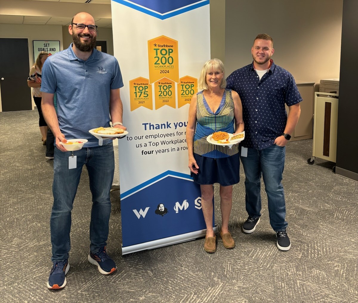 Three Michigan Millers employees standing in front of Top Workplaces celebration poster.